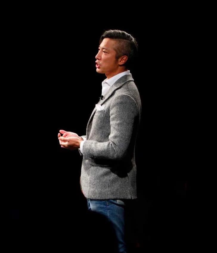 黒い舞台に立ち、手を組んで語りかける秦卓民の全身