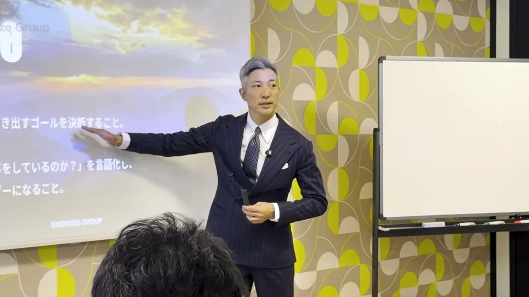 研修室でスクリーンを指しながら講義する紺のスーツ姿の秦卓民。右にホワイトボード、手前に受講者