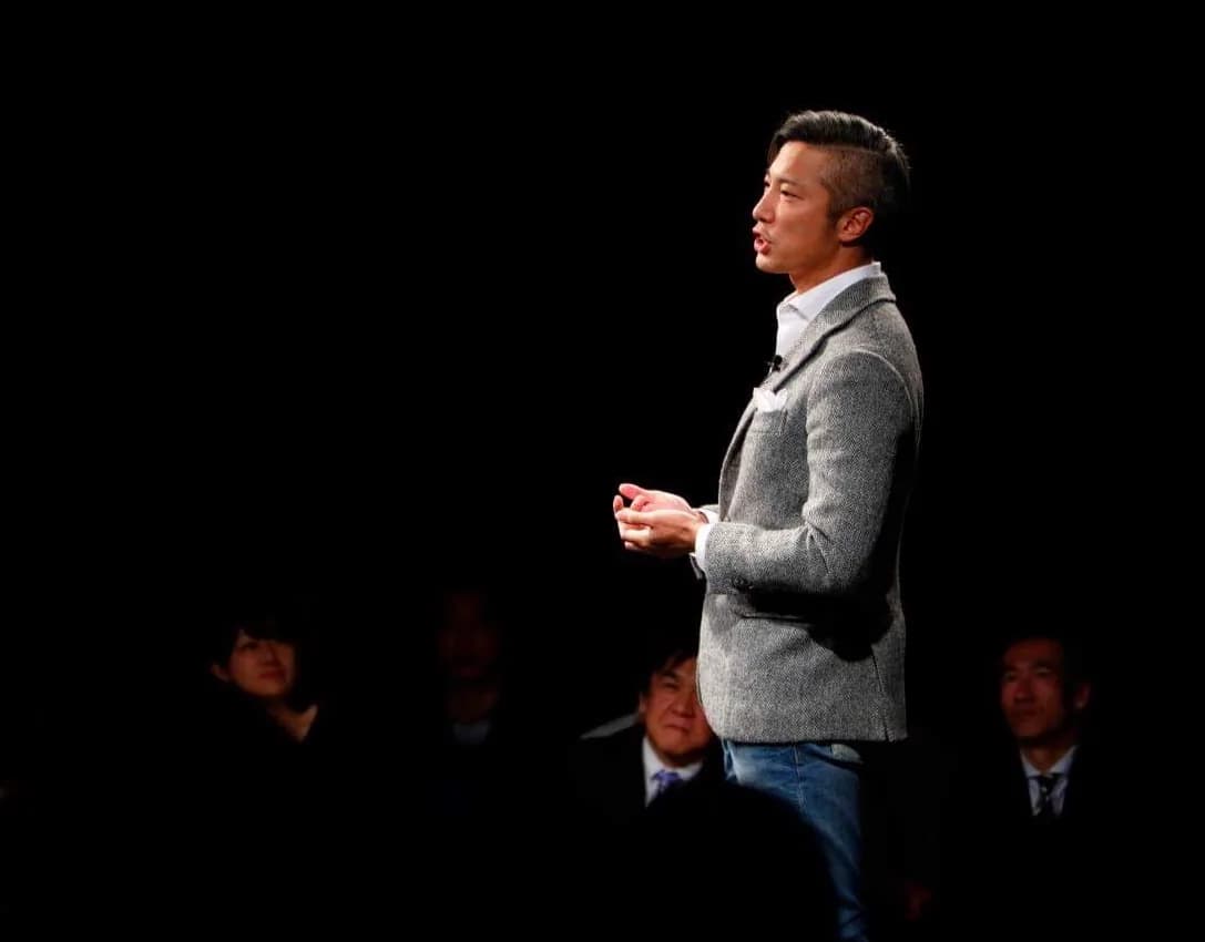 暗い会場の壇上で聴衆に語りかける秦卓民。手前に聴講する参加者が並ぶ